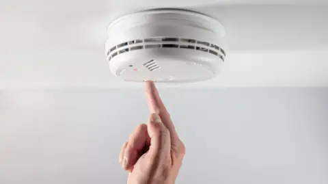 Getty Images/Brian A Jackson A white smoke alarm attached to a white ceiling.  A man's hand is partially visible as he presses a button on the alarm during a test.