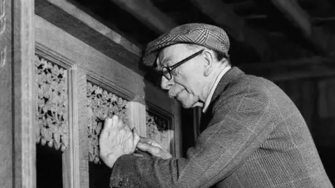 Alamy A black and white image of a man in a tweed outfit carving wooden church altars.