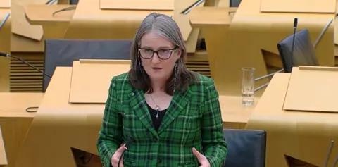 Scottish Parliament Maree Todd wears a green tartan jacket, black top, silver necklace and black-framed glasses with a retainer chain. She is standing in the chamber in Holyrood, with wooden desks and black seats behind her.