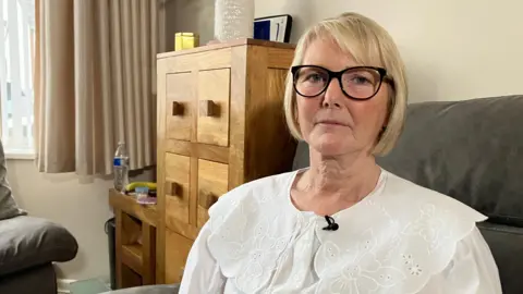 Julie Morton, who has a blonde bob and large black framed glasses wears a vintage style large collared white top. She is sitting on a grey sofa and a wooden cupboard behind her. 