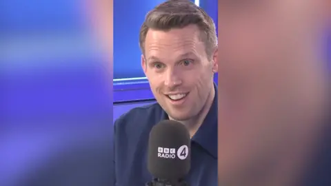 A man with short, light brown hair smiles as he talks into a microphone.