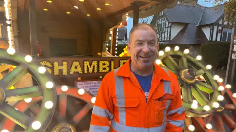 A man in orange hi-vis standing in front of a carnival cart reading 'Shambles' with wooden wheels covered in light bulbs on the front