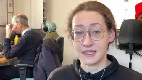 Hari Reed has medium length dark hair which is tied up, and glasses. She is in an office, with a colleague working at a desk behind her.