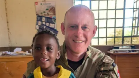 Mr McMahon is sat in a room in Kenya with a young child on his lap. He is wearing Army fatigues and has a shaved head and is smiling at the camera. The child has a blue and yellow top on. There is a window behind them and some desks or tables with paperwork on.