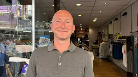 Man with bald head stands outside a shop with clothing on hangers