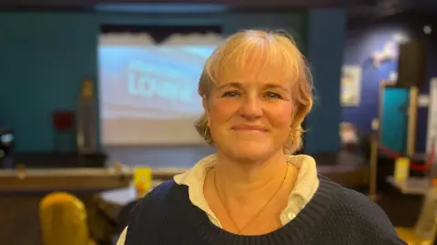 A lady smiling at the camera wearing a white blouse with a blue jumper. She is smiling at the camera. She is in a bar which is out of focus in the background.