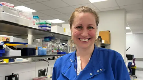 University Hospital Southampton Dr Jemma Longley is in a laboratory and looking at the camera smiling. She wears a blue lab coat and has her hair swept back
