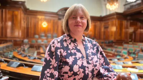 Aisha Iqbal/BBC A woman in a floral pink and blank dress. She has blonde, bobbed hair. She stands in a council chamber which is otherwise empty.