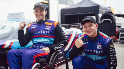 Dan Bathie A man using a wheelchair next to a younger man who is crouching. They are both smiling, and are wearing race suits. They are both in front of one of their team cars. 