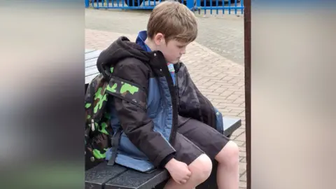 Victoria Dean Alex in his school uniform sat on a bench, with his rucksack on his back. He is pictured side-on and he is looking at the ground. He looks sad.
