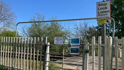 Tom Ingall/BBC A large metal fence stands in front of a gate by a railway level crossing. On the fence is a note saying CROSSING CURRENTLY OUT OF USE. To one side is a STOP LOOK LISTEN warning sign. In the background are trees and a blue sky.
