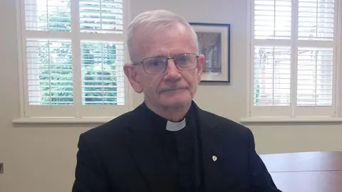 Bishop Alan McGuckian is looking directly to the camera. He has short grey hair and is wearing a black blazer and black shirt with a white priest collar. 
