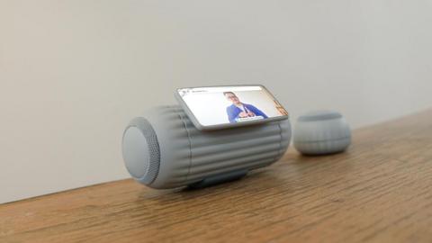 The grey speaker sitting on a wooden table. The main speaker has rounded edges and a screen in the middle of it about the size of a smartphone and next to it is a smaller, round speaker
