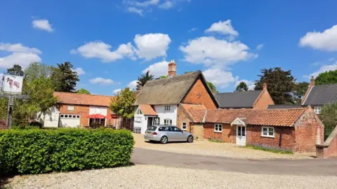 Ryan Townsend A pub with a thatched roof and white-painted bricks with a driveway lined by hedgerows in the foreground