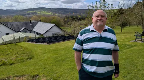 A person standing on a grassy slope overlooking houses and rolling hills under a partly cloudy sky. Bob Horton is shown in the foreground wearing a striped top, with the Carmarthenshire landscape and buildings behind him.