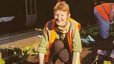 Edwina Simpson potting plants at a train station. She has red hair and is wearing a fluorescent orange jacket.