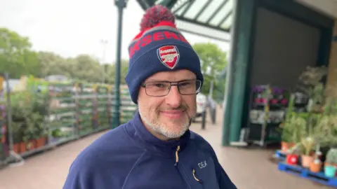 A man with a blue fleece and blue Arsenal woolly hat stands in a garden centre. He wears glasses and has a grey beard and smiles at the camera.