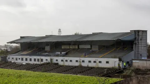 a dilapidated looking seating stand. Weeds are growing from between the seats