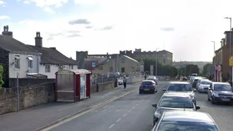 Legrams Lane in Bradford, looking towards a bus shelter and a row of houses. There is queuing traffic on the road, with cars also parked up at the side. 