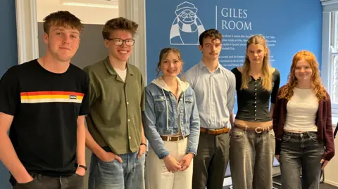 Jon Wright/BBC Six young people stand in a line, smiling at the camera. The wall behind has the words Giles Room.