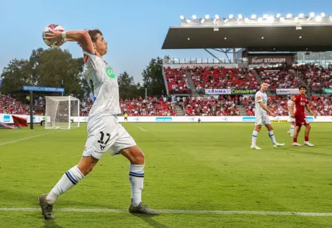 Getty Images Callan Elliot in a white football strip takes a long throw-in as a couple of other footballers look on with a stadium and floodlights in the background