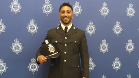 Rehaan Akhtar in Police uniform, holding his constable's hat. He is standing in front of a board covered in South Wales Police logos. This is shaped like a seven pointed star with a crown atop, with the Prince of Wales' feathers arranged in the middle