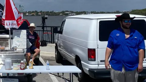 Bernd Debusmann Jr/BBC News Dirk Fraser, dressed in blue and wearing a black cowboy hat, is pictured on a bridge next to his hot dog stand with the waterways of Palm Beach in the background.