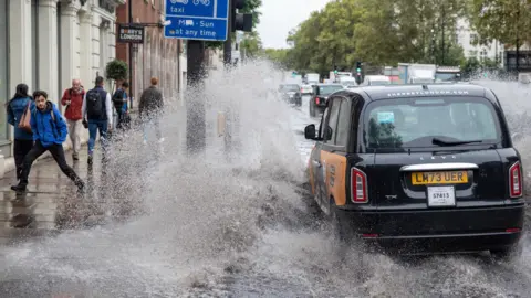 Simon Lamrock A black taxi pulls over into a large puddle, splashing people walking past on the pavement. 
