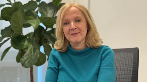 Helena Mangan, Head of Work and Family at Employment and Social Security for the States of Jersey wearing a teal long‑sleeved top sits in an office chair next to a large potted plant. The setting appears to be a bright indoor office space with light‑coloured walls and a window in the background.