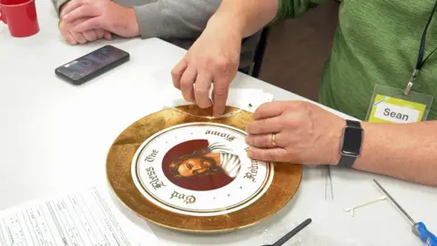 Noticias de la BBC. Sobre la mesa hay una placa con el nombre de una familia. Tiene un amplio borde dorado. En el centro hay una imagen de Jesús sobre un fondo rojo. Una inscripción dorada alrededor de Jesús dice: «Dios bendiga el Hogar Robinson». Un reparador con la placa, Sean, tiene las manos sobre la placa, tocando una gran grieta. Lleva una camiseta verde.