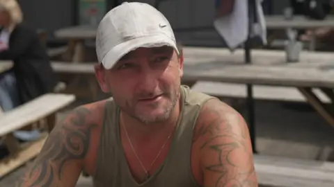 BBC Jodie is wearing a white baseball cap, a khaki coloured vest and a very thin silver-coloured necklace. He has a stubbly beard in light brown and grey colours and tattoos along both arms. He is sat on a wooden bench and behind him are four sets of table and benches with a parasol in the middle.