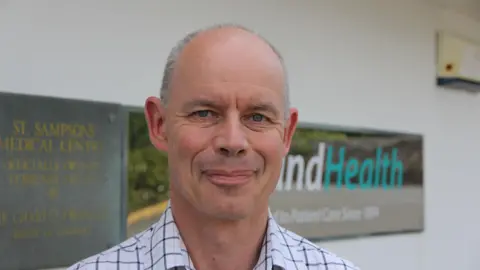 A mostly bald man, with short grey hair on the sides smiles at the camera. He had blue eyes and is wearing a blue checked shirt. Behind him, a shiny sign reads 'IslandHealth', and a plaque says 'St Sampson's Medical Centre.2