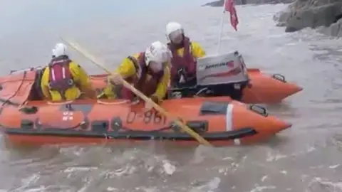Three lifeboat crew members in yellow, all-in-one uniforms and white helmets, in a red dingy.