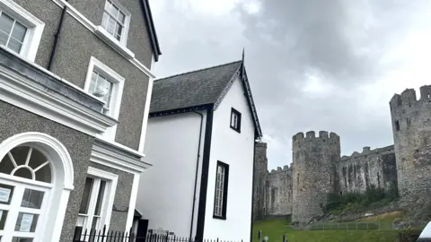 A grey-brown pebbledashed 18th century building with white smooth render around the windows and a slate plaque marked "Bodreinallt Surgery" occupies the left hand side of the photo. It has a porch with an arched window and NHS notices stuck in some of the panels. On the right is a partial view of Conwy's medieval castle with its round towers. The gable end of a white building with black smooth window surrounds is in the centre.
