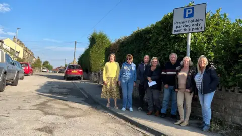 Seven middle-aged people - five women and two men, all white and aged in their 50s or so - standing below a 'permit holders parking only' sign. Behind them is a street scene showing parked cars and a row of houses with neatly trimmed hedges and trees. The road itself looks slightly rough and has several potholes.