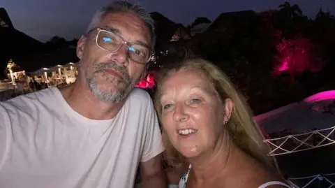 A couple, both about 60 years old, standing outside in the dark, with lights visible in the background. The man on the left has short grey hair and a matching beard and is wearing glasses and a pale pink T-shirt. The woman on the right has long blonde hair and is wearing ear studs and a white top.