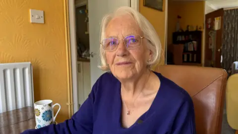 BBC Lorraine Boyce is an elderly woman. She is sitting in her lounge on a brown chair. We can see a bookcase behind her. There is a mug on the table next to her. She is wearing clear thin-rimmed glasses and a dark blue jumper.