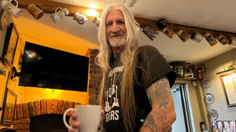 Rick with long grey hair before getting it cut off. He is holding a white mug and is standing to the side in a pub looking into the camera.