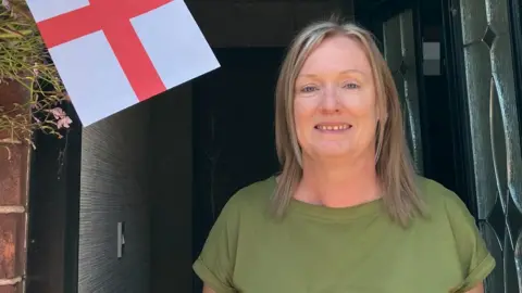 Maxine Simeone, who has shoulder length blonde hair and a green top, smiles at the camera from inside the doorway of a terraced house. A red and white England flag is to her right. 