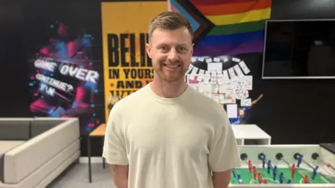 Mark Ansell/BBC Deputy manager of the youth centre, Tom Chambers, smiling at the camera. He is inside the Epic Youth Hub and has a cream t-shirt on and is standing in front of a table football game.