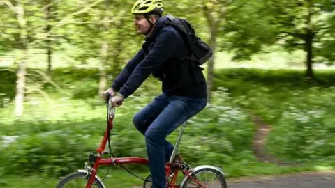 A man wearing a yellow bike helmet rides a red city bike. The background is trees and shrubbery but is blurred because of the travelling cyclist. 