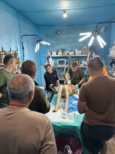 Vīrietis, kurš valkā tikai šortus, guļ uz gultas, ko ieskauj seši citi vīrieši. Uz sienām ir plaukti ar dažādiem medicīniskiem priekšmetiem, un medicīnas priekšmeti balstās arī uz gultas blakus vīrietim.