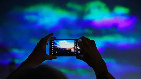 Reuters person takes a picture on their mobile of people watching the night show with strand of purple, blue and green in the sky backdrop