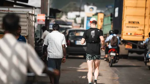 James Poole Mr Robinson running alongside traffic on a road in India. Beside him are large lorries, cars, trailers and mopeds. Mr Robinson is wearing green and black patterned shorts, a black t-shirt, running vest, and red and white cap.