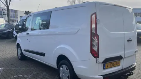 Family A white van, with one side and the back visible, dominates the photo. Parts of other vehicles and buildings are in the background.