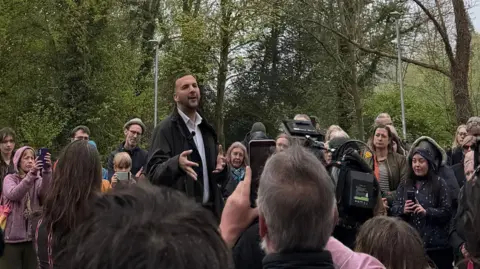 Zack Polanski wearing a dark anorak addressing a crowd of people with their phones and cameras raised
