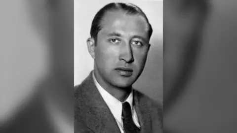 A black and white image of Duško Popov wearing a suite jacket over a white shirt and dark tie. He has short slicked-back hair.