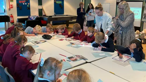 BBC A class room of children sat around a large table. There are a few teachers stood around the table.