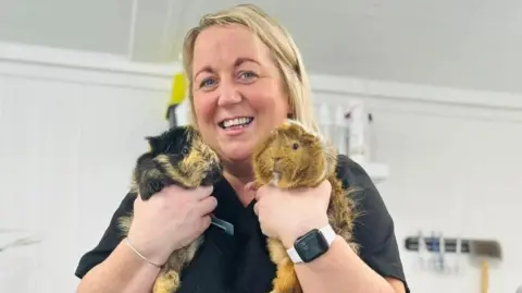 Claire McConaghy. Claire McConaghy. She has blonde hair and blue eyes and wearing dark coloured scrubs, an Apple watch one her left wrist and silver bracelet on her right. Claire is holding up two guinea pigs, the one on the left is black and tan and the one on the right is mainly tan with bits on white.