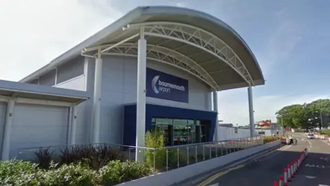 Google Google Street View of the entrance to Bournemouth airport terminal building. It is a large building clad with white panels and with a large arched portico sticking out over the footpath below. The Bournemouth Airport sign above the glass doors is Navy blue with white writing. The road in front has cones down the middle and a man wearing hi-viz can be seen in the distance.